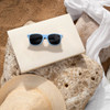 Light blue RPET sunglasses rest on a beige surface, surrounded by natural stone and a straw hat on sand.