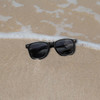 Classic black sunglasses resting on a sandy beach, with gentle waves in the background.