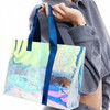 A transparent PVC tote bag with a colourful iridescent surface and navy blue handles, held by a person.