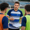 A V-neck raglan tee shirt in blue and white, featuring a logo, worn by a coach discussing with players on a field.