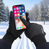 Black touchscreen winter gloves are worn while using a smartphone in a snowy outdoor setting with pine trees.
