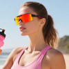 Frameless sports sunglasses with mirrored red lenses and black accents, held by a woman in a pink top, near a beach.