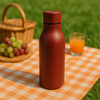 A drink bottle in a matte red finish, with a sleek design and a logo, placed on a picnic blanket.