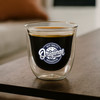 A double wall glass featuring a black beverage and a logo, set on a wooden table. The glass has a transparent design.