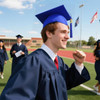 A young man wearing a blue mortarboard graduation cap and navy gown smiles, celebrating on a sports field.