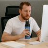 A drink bottle in blue and black, featuring a logo, is held by a person seated at a desk with a computer.