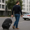 A wheeled duffel bag in black, being pulled by a man wearing a navy sweater and jeans on a city street.