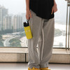 A drink bottle in vibrant yellow, featuring a carry handle, held by a person in casual grey pants. The background shows a city view.