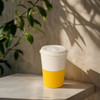 A drink bottle in white and yellow, featuring ribbed sides and a lid, set against a light background with plant shadows.