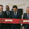 A red ribbon marked "BOXING" with a logo, held by four men in suits, in a setting with a "Congratulations" banner.