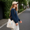A woman carries a cream vintage style canvas bag with a brown strap, wearing a navy sweater and a beige cap.
