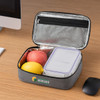 A grey insulated lunch bag opened to reveal two apples, a lemon, and a food container inside. It features a logo.