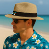 A straw Panama beach hat in tan with a black band, worn by a man in sunglasses and a floral shirt.