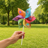A hand holds a colourful six-leaf pinwheel with various designs, set against a green park backdrop.