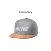 A grey snapback cap with a cork brim, featuring embroidery on the front and a logo displayed.