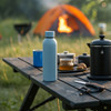 A drink bottle in light blue on a wooden table, surrounded by camping gear and a tent in the background.