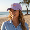 A lilac baseball cap with a logo, worn by a smiling woman on a beach, with a scenic ocean view in the background.