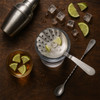 A stainless steel bar strainer with a sleek design, accompanied by lime wedges, ice cubes, and a cocktail shaker.