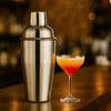 A stainless steel cocktail shaker in a polished finish, next to a vibrant cocktail in a glass.