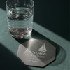 An octagonal metal coaster with a silver finish rests on a green surface beside a glass of water.