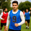 A blue training bib with a number 9, featuring a logo, worn over a long-sleeved shirt on a grassy field.