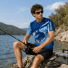A blue polyester spandex fishing T-shirt with a fish graphic and logo, worn by a man at a lakeside.