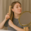 A bamboo comb being held by a person with straight hair, facing a round mirror. The comb is light brown.