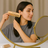 A fine-tooth bamboo small comb in beige held by a person in front of a circular mirror.