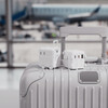 A white travel plug sits on a silver suitcase, with an airport background featuring blurred planes and terminals.