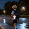 A reflective keychain in the shape of a person hangs from a child's backpack as he walks on a wet street at night.