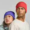 A red and a purple beanie are worn by a man and a woman, featuring a ribbed texture and a branding logo.