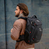 A man with long hair wearing a brown jacket carries a black Swissdigital Neptune Backpack. The backpack has a logo.