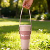 A drink bottle holder in soft pink with a strap, resting on green grass, featuring a logo on the front.