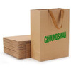 A brown recycled paper bag with a paper webbed handle, next to a stack of flat paper bags.