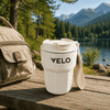 A white double wall coffee cup with a lid and a strap, placed on a wooden table near a forested lake.