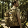 A tactical backpack in a tan colour, equipped with multiple straps, is worn by a man in a forested setting.