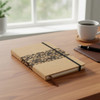 A cork notebook featuring a black design, resting on a wooden desk with a coffee cup and pen in the background.