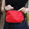 A red bum bag with a black zip, held by a person in a black top, featuring a logo.