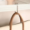 A silver foldable desk hook is attached to a shelf, holding a brown-handled bag. The background is light-coloured.