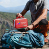A red empty first aid kit bag with a handle, placed on a blue backpack outdoors. The bag features a logo.