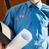 A man in a blue shirt holds rolled black and white papers, featuring a metallic name badge on his chest.