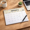A horizontal wall calendar for January 2026 with a floral design, placed on a wooden table alongside a coffee mug and plant.