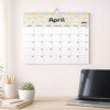 A wall calendar featuring a floral design and a section for April 2027, displayed with a plant and a laptop.