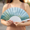 A fabric hand-held fan featuring a gradient design in soft blue and green tones, held by a person.