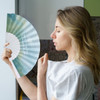 A fabric hand-held fan featuring a gradient design in soft blue and green colours, being held by a woman with blonde hair.