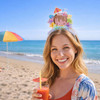 A woman at the beach wears a colourful headband with a decorative topper while holding a drink.