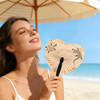 A heart-shaped plastic paper fan with designs, held by a smiling woman at a beach under an umbrella.