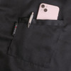 A black half apron made of 100% polyester featuring a pocket, holding a pen and a smartphone.