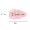 A teardrop-shaped, iridescent hair clip in pink and gold with a logo, measuring 5 cm by 2.7 cm.