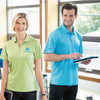 A green women's polo shirt and a blue men's polo shirt, both featuring a logo, displayed in a bright office setting.